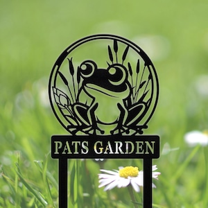 Puede incluir: Estaca de jardín de metal negro con un diseño de rana dentro de un marco circular, rodeada de juncos. La estaca tiene el texto "PATS GARDEN" y está colocada sobre un fondo de hierba verde y margaritas blancas.