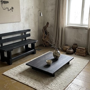 Peut inclure: Aménagement de salon minimaliste avec une table basse en bois foncé, un tapis texturé et un banc noir. Un vase avec des branches et un petit bol sont posés sur la table. La pièce présente des tons neutres et une lumière naturelle.