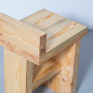May include: A handcrafted wooden stool with a natural finish. The stool features a simple, geometric design with a rectangular seat and a backrest. The wood grain is visible, adding to the rustic aesthetic. The stool measures approximately 18 inches tall.
