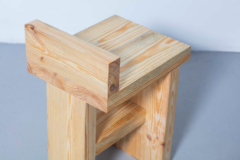 May include: A handcrafted wooden stool with a natural finish. The stool features a simple, geometric design with a rectangular seat and a backrest. The wood grain is visible, adding to the rustic aesthetic. The stool measures approximately 18 inches tall.