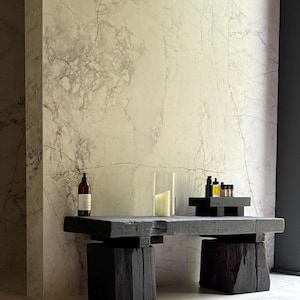 May include: A dark, rustic wooden table with a rectangular top and block legs. The table holds a glass candle holder with a lit candle, a bottle, and small containers. The background is a marble wall.