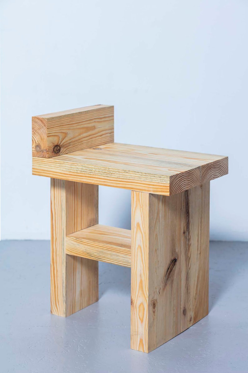 May include: A small, handcrafted wooden stool with a natural finish. The stool features a rectangular seat, a backrest, and a lower shelf. The dimensions are approximately 12 inches wide, 12 inches deep, and 24 inches high.