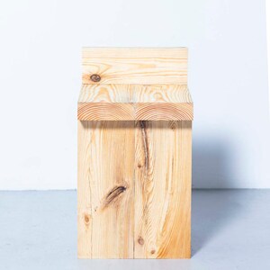 May include: A natural wood stool with a rectangular seat and a solid, block-like base. The wood grain is visible, and the stool is constructed from several pieces of light-colored lumber. The design is simple and modern.