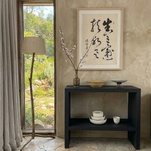 May include: A black wooden console table with two shelves, holding a bronze vase with flowering branches, a yellow bowl, and a patterned dish. Below, three stacked white bowls and a small cup sit. A framed calligraphy print hangs above.