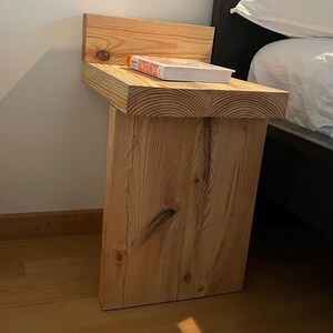 May include: A rustic, wooden bedside table with a natural finish. The table features a thick, rectangular top and a single, sturdy leg. A book with a colorful cover rests on the tabletop. The table is positioned next to a bed with white bedding.