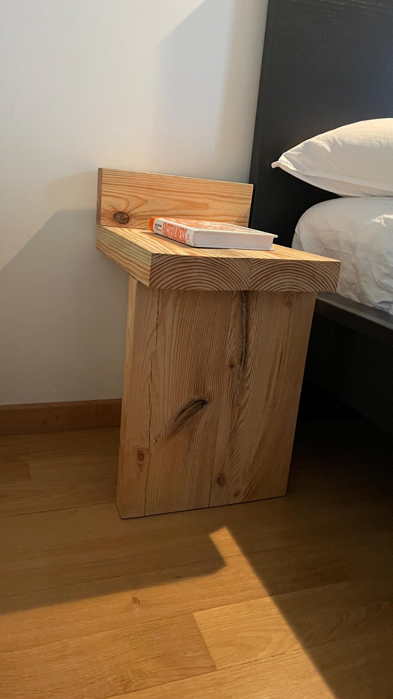 May include: A rustic, wooden bedside table with a natural finish. The table features a thick, rectangular top and a single, sturdy leg. A book with a colorful cover rests on the tabletop. The table is positioned next to a bed with white bedding.