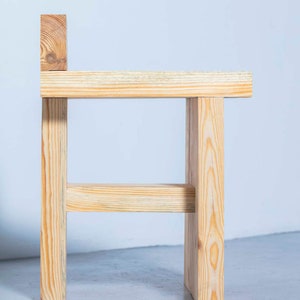 May include: A small, handcrafted wooden stool with a simple design. The stool is made from light-colored wood, featuring a rectangular seat, four legs, and a small backrest. The wood grain is visible, adding to its natural aesthetic.