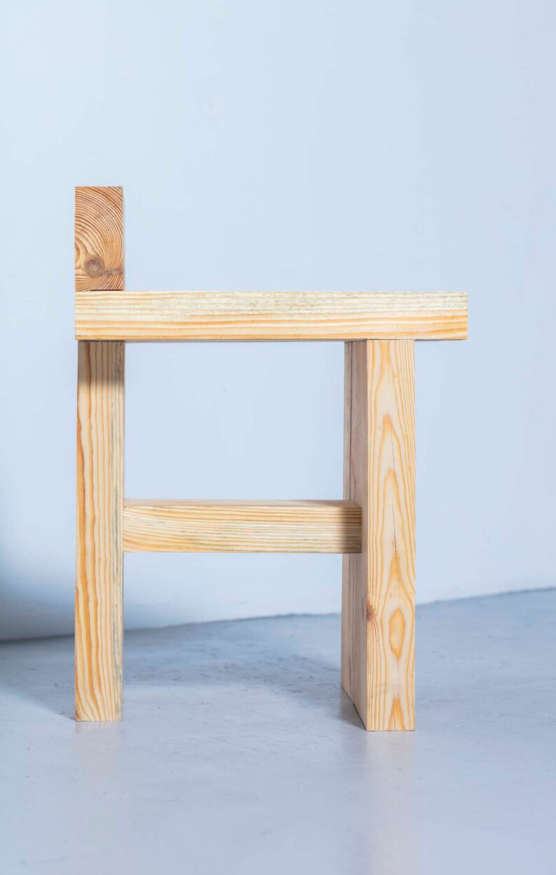 May include: A small, handcrafted wooden stool with a simple design. The stool is made from light-colored wood, featuring a rectangular seat, four legs, and a small backrest. The wood grain is visible, adding to its natural aesthetic.