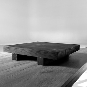 Op de afbeelding: Een zwart houten platform met een vierkant bovenblad en vier poten. Het platform is gemaakt van ruw hout en heeft een rustieke uitstraling.