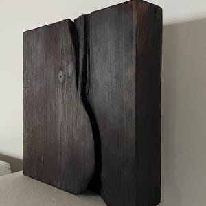 Könnte beinhalten: Eine dunkelbraune, rechteckige Holzskulptur mit einem vertikalen Spalt in der Mitte. Die Holzmaserung ist sichtbar und die Skulptur hat eine raue, strukturierte Oberfläche. Das Stück wird auf einer weißen Oberfläche präsentiert.