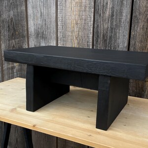 Könnte beinhalten: Ein schwarzer Holz-Couchtisch mit einer rechteckigen Tischplatte und zwei quadratischen Beinen auf jeder Seite. Der Tisch ist aus recyceltem Holz gefertigt und hat eine rustikale Oberfläche.