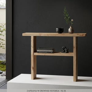 May include: A light-colored wooden console table with a lower shelf, holding books and a decorative sculpture. The top shelf displays a black bowl, a small vase with greenery, and a small vase with a flower. The text "ZAPISANA W DREWNIE HISTORIA - Nr. 01" is at the bottom.