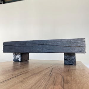 May include: A dark grey wooden shelf, supported by two smaller wooden blocks. The shelf has a textured, weathered appearance. The blocks are painted white and grey. The shelf is displayed on a wooden surface.