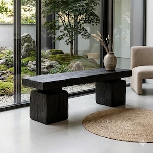 Könnte beinhalten: Eine schwarze Holzbank mit rechteckiger Oberseite und zwei quadratischen Stützen. Eine beige Vase mit getrockneten Gräsern steht auf der Bank. Ein runder, gewebter Jute-Teppich liegt auf dem Boden.