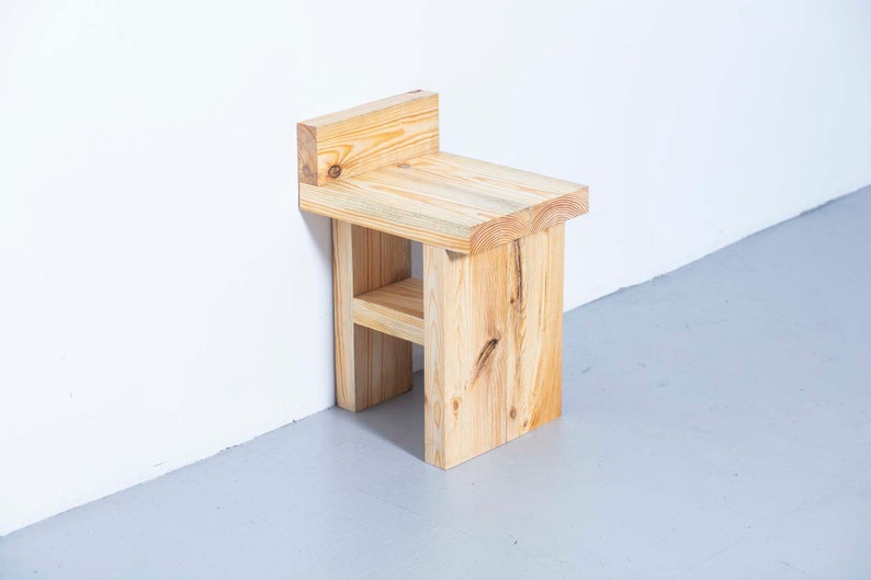 May include: A small, natural wood stool with a backrest. The stool is constructed from light-colored, unfinished lumber. The design features a rectangular seat, a back support, and a lower shelf. The stool is approximately 12 inches wide, 10 inches deep, and 18 inches tall.