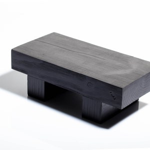 Puede incluir: Un taburete de madera negro con una parte superior rectangular y cuatro patas. El taburete está hecho de madera maciza y tiene un acabado natural.