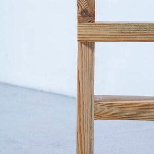 May include: A close-up of a wooden chair with a natural finish. The chair has a simple design with a vertical support and horizontal slats. The wood grain is visible, and the chair appears to be made of pine.