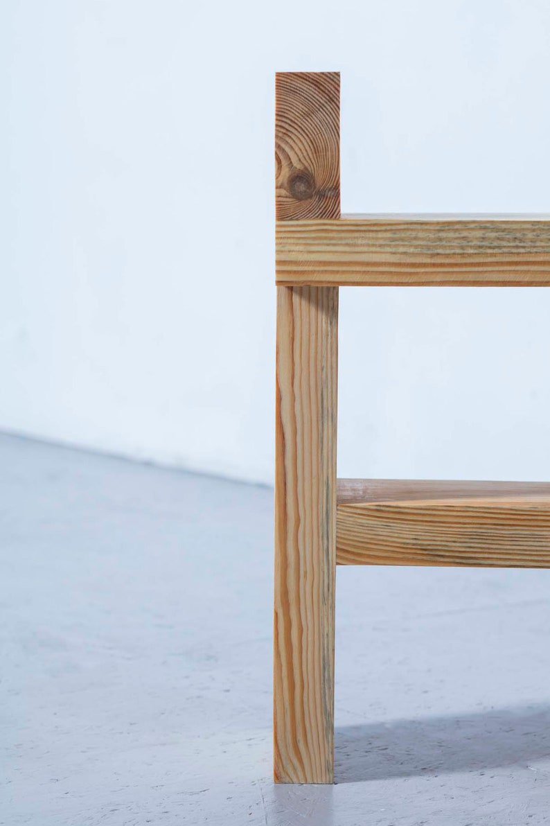 May include: A close-up of a wooden chair with a natural finish. The chair has a simple design with a vertical support and horizontal slats. The wood grain is visible, and the chair appears to be made of pine.