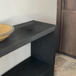Peut inclure: Une console en bois noir avec un plateau rectangulaire et une étagère inférieure, présentant une texture de grain de bois visible. Un bol peu profond, rond et de couleur beige avec un bord foncé repose sur le côté gauche de la table.