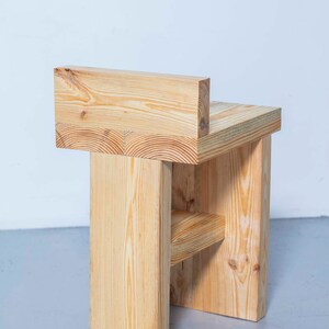 May include: A handcrafted wooden chair with a minimalist design. The chair is constructed from light-colored wood, featuring a rectangular seat, backrest, and supporting legs. The natural wood grain is visible, adding to its rustic charm. The chair's dimensions are approximately 18 inches wide, 16 inches deep, and 30 inches high.