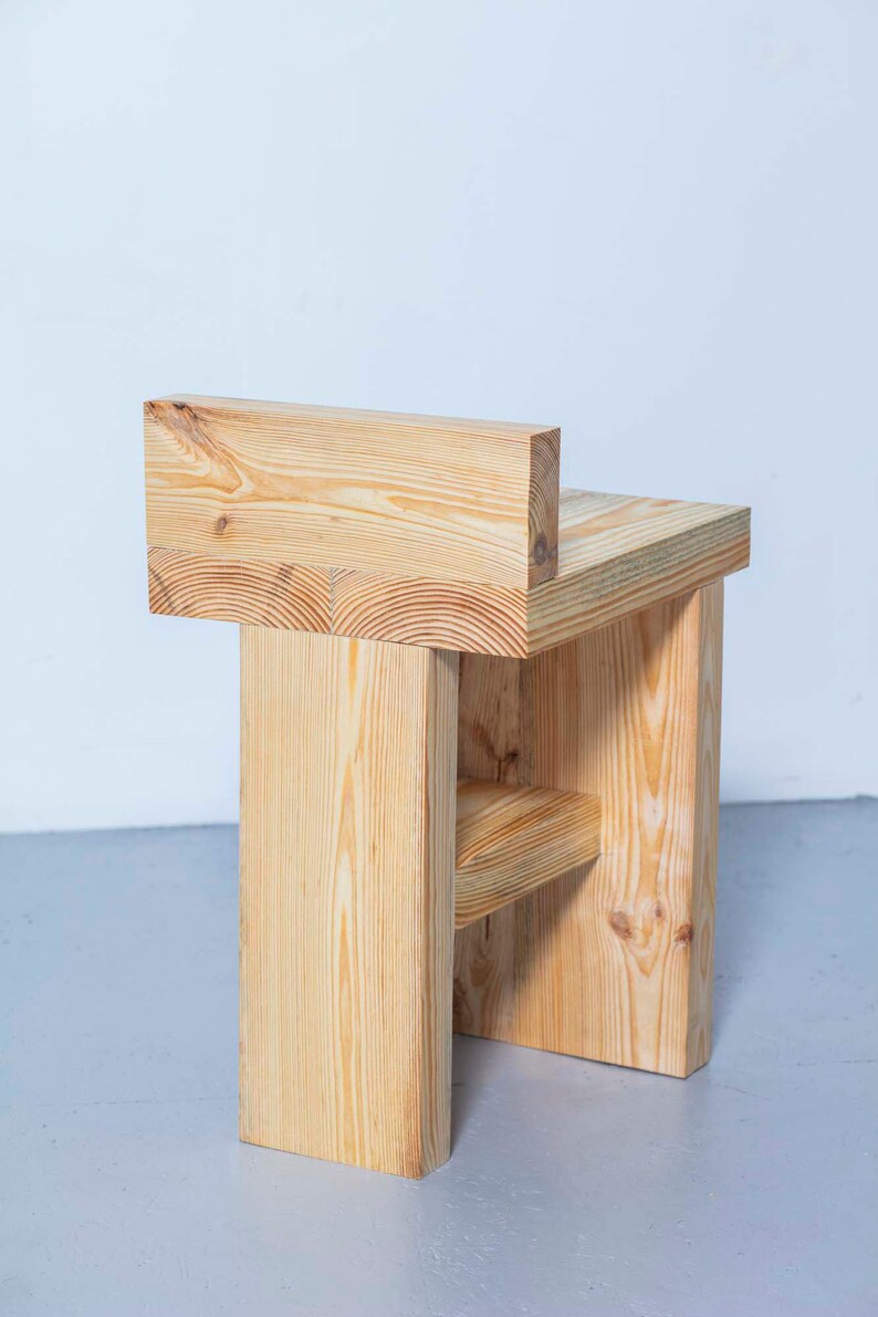 May include: A handcrafted wooden chair with a minimalist design. The chair is constructed from light-colored wood, featuring a rectangular seat, backrest, and supporting legs. The natural wood grain is visible, adding to its rustic charm. The chair's dimensions are approximately 18 inches wide, 16 inches deep, and 30 inches high.