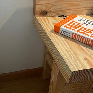May include: A wooden side table with a book titled "Factfulness" on top. The table is made of light-colored wood and has a simple, sturdy design. The book has an orange cover with white and black text.