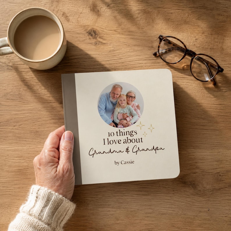 Hand holding a personalized “10 Things I Love About Grandma & Grandpa” photo board book on a wooden table beside a cup and glasses. Cream cover with a circular family photograph.