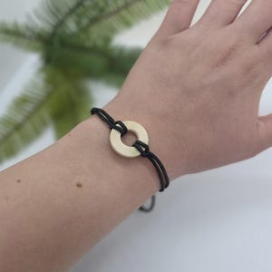 May include: A minimalist bracelet featuring a cream-colored circular pendant and black cord. The bracelet is tied with a sliding knot closure. The jewelry is simple and elegant, suitable for everyday wear.