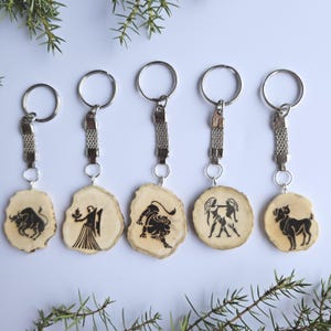 May include: Five wooden keychains with silver metal rings and chains. Each keychain features a different black zodiac symbol burned into a light-colored wood slice. The keychains are arranged on a white surface with green sprigs.