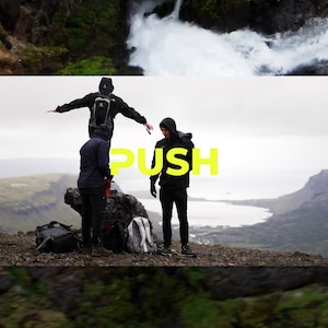 May include: Three people stand on a rocky cliff overlooking a body of water. The person in the middle is wearing a black jacket and has the word "PUSH" written in yellow letters on their chest. The sky is overcast and the water is a light blue color.