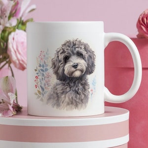 Könnte beinhalten: Weiße Keramiktasse mit einer Aquarellillustration eines grau-schwarzen Hundes und floralen Akzenten. Die Tasse steht auf einer rosa Geschenkbox, mit rosa Blumen und einer Rose im Hintergrund. Die Tasse hat einen gebogenen Henkel.
