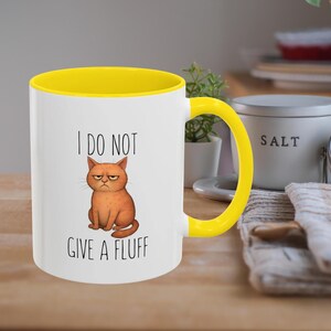 Puede incluir: Taza de cerámica blanca con interior y asa amarillos. La taza presenta una ilustración de un gato naranja gruñón y el texto "I DO NOT GIVE A FLUFF". La taza está sobre una superficie de madera.
