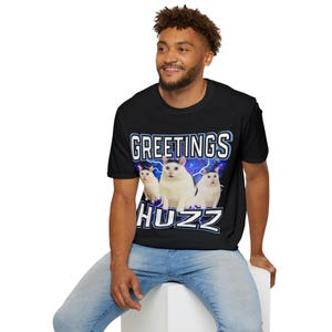 Op de afbeelding: Zwart T-shirt met een afbeelding van drie witte katten en de tekst "GREETINGS HUZZ" in blauw en wit. De katten staan tegen een blauwe bliksemachtergrond. Het T-shirt wordt door een persoon gedragen.