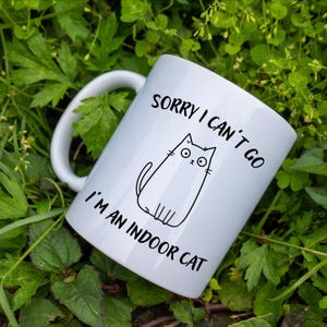 Puede incluir: Taza de cerámica blanca con un dibujo de contorno negro de un gato y el texto "SORRY I CAN'T GO I'M AN INDOOR CAT". La taza tiene un asa curva y está rodeada de follaje verde.