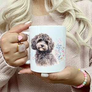 May include: White ceramic mug featuring a watercolor illustration of a gray and brown dog with a floral background. The mug is held by a person wearing a pink sweater and rings. The mug is a standard size for coffee or tea.