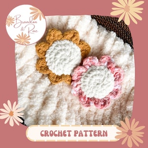 Può includere: Modello a uncinetto per una coperta bianca e soffice con due grandi fiori all'uncinetto, uno in giallo senape e uno in rosa. Il modello è intitolato "Brandon & Rae Crochet Pattern".