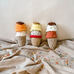 Peut inclure: Trois c&ocirc;nes de cr&egrave;me glac&eacute;e en crochet avec des boules de cr&egrave;me glac&eacute;e de diff&eacute;rentes couleurs. Les c&ocirc;nes sont pos&eacute;s sur une surface en tissu beige avec des p&eacute;pites color&eacute;es &eacute;parpill&eacute;es autour d'eux.