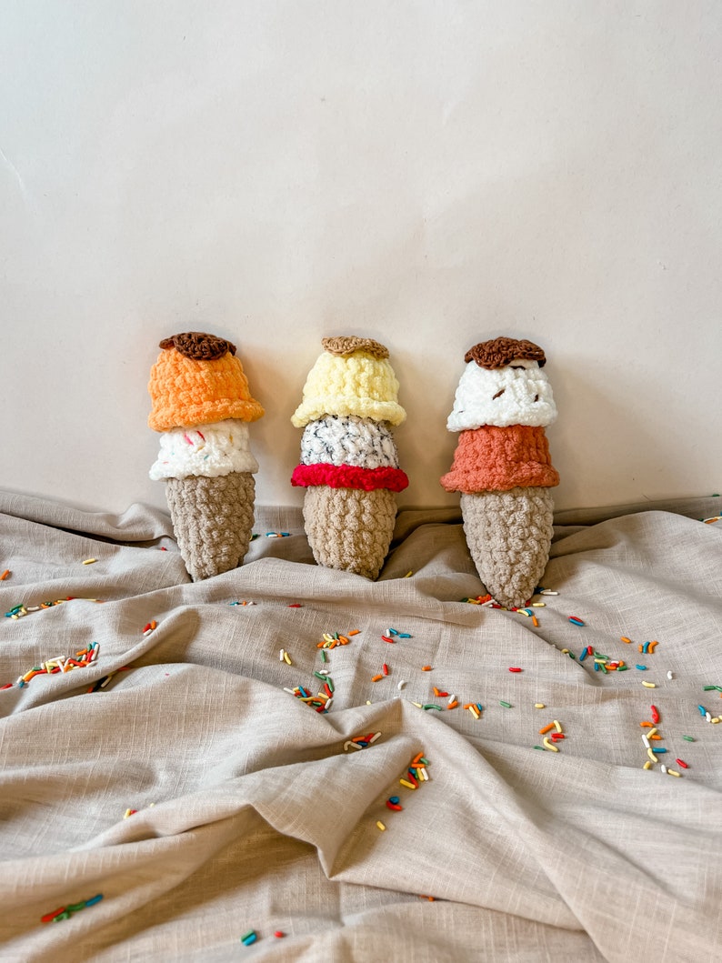 Peut inclure: Trois c&ocirc;nes de cr&egrave;me glac&eacute;e en crochet avec des boules de cr&egrave;me glac&eacute;e de diff&eacute;rentes couleurs. Les c&ocirc;nes sont pos&eacute;s sur une surface en tissu beige avec des p&eacute;pites color&eacute;es &eacute;parpill&eacute;es autour d'eux.