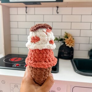Peut inclure: Un jouet en forme de cornet de glace en crochet, r&eacute;alis&eacute; avec du fil brun, rose et blanc. Le cornet est tenu dans une main devant une cuisine jouet.