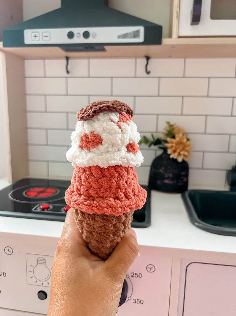 Peut inclure: Un jouet en forme de cornet de glace en crochet, r&eacute;alis&eacute; avec du fil brun, rose et blanc. Le cornet est tenu dans une main devant une cuisine jouet.