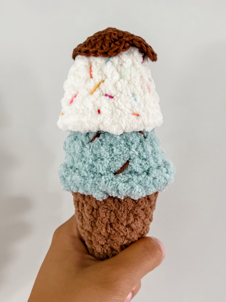 Peut inclure: Un jouet en forme de cornet de glace crochet&eacute; avec trois boules de glace. La boule du haut est blanche avec des p&eacute;pites color&eacute;es, la boule du milieu est bleu clair et la boule du bas est brune.