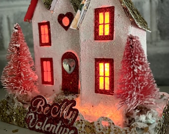May include: A white papercraft house with red accents and a red heart door. The house has lit windows and is decorated with glitter. The house is sitting on a base with a gold glitter finish and the words "Be My Valentine" in red letters.