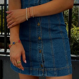 May include: A denim dress with a button-down front and a fitted silhouette. The dress is paired with a gold ring and several bracelets, including a gold bracelet with red, blue, and white flower accents and beaded bracelets.