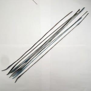 May include: A collection of thin, dark-colored, elongated objects, possibly stems or wires, arranged diagonally against a white background. Some have a slight blue-green hue at the tips, suggesting a natural or treated material. The objects are of varying lengths.
