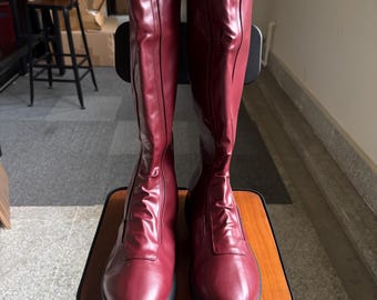 May include: A pair of tall, burgundy-colored boots. The boots are made of a smooth material and have a knee-high design. They are placed on a small wooden stool with a black metal frame. The boots have a sleek, modern appearance.
