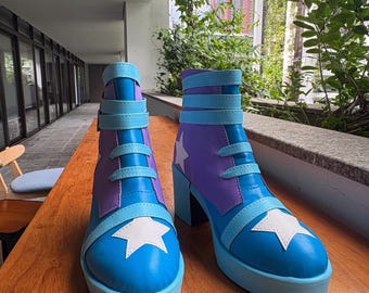 May include: A pair of ankle boots featuring a vibrant color scheme of bright blue, purple, and white. The boots have a thick platform sole, a block heel, and multiple horizontal straps across the front. Each boot is decorated with a large white star.