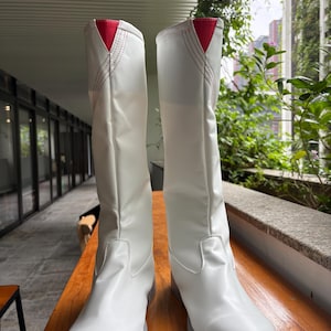 May include: A pair of knee-high white boots featuring a smooth synthetic finish, a rounded toe, and a flat grey sole. Each boot has a distinctive V-shaped cutout at the top centre with a bright red triangular accent and decorative red stitching.