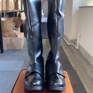 May include: A pair of black knee-high boots with a glossy finish. The boots have a strap detail across the top of the foot and a buckle on the side. They are displayed on a small wooden stool.