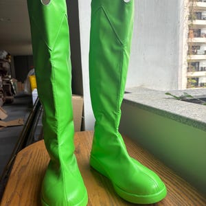Puede incluir: Un par de botas altas de color verde brillante. Las botas tienen una textura lisa y brillante y una punta redondeada. La caña de las botas es alta, con un ligero corte en forma de V en la parte superior. Las botas están sobre una superficie de madera.