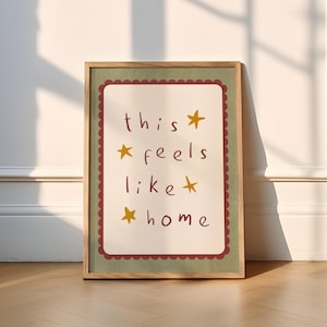 K&ouml;nnte beinhalten: Ein gerahmter Druck mit gr&uuml;nem Hintergrund und rotem Rahmen. Der Druck zeigt den Text "this feels like home" mit goldenen Sternen.
